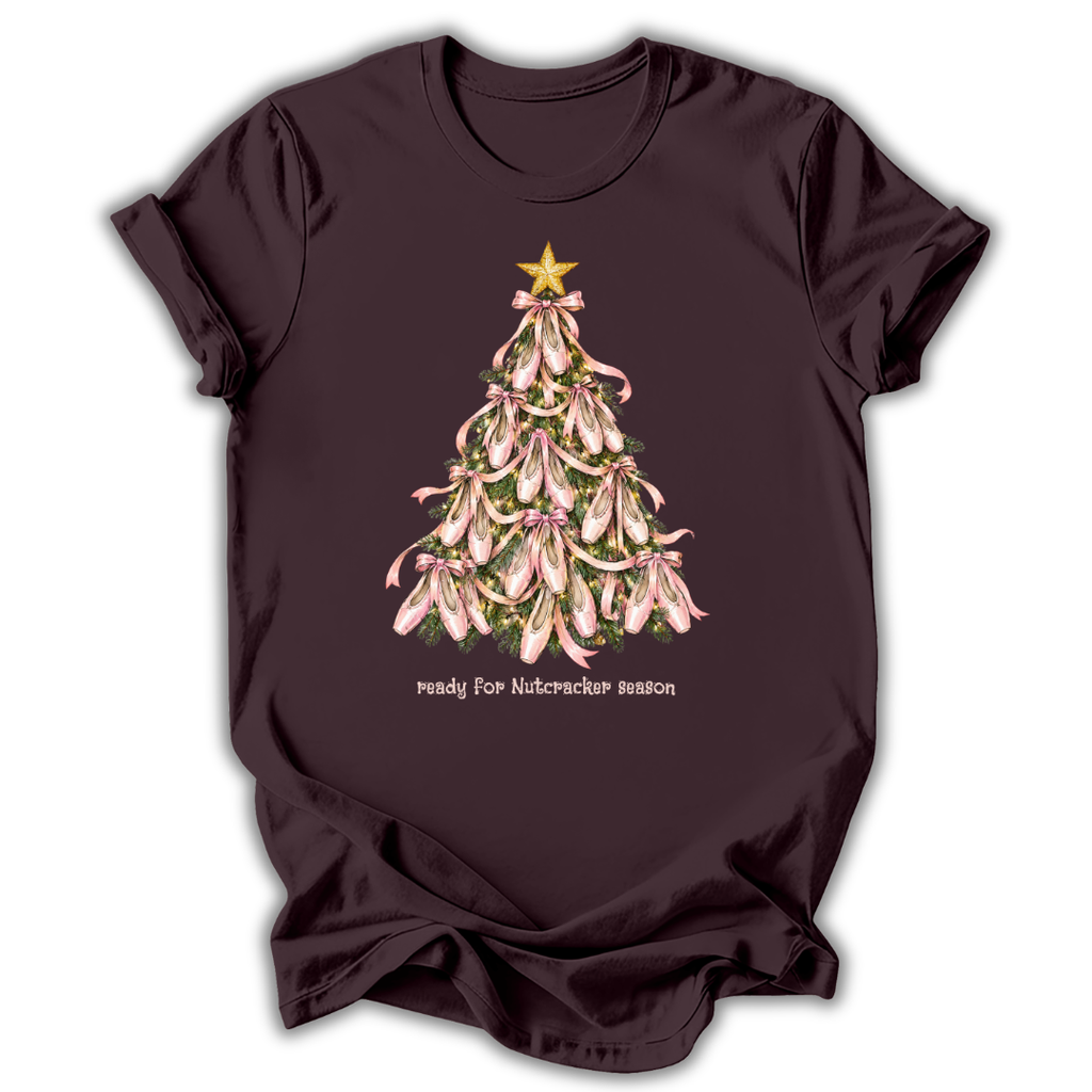 A dark plum-colored short-sleeve t-shirt with a graphic of a Christmas tree made of pink lilies with a gold star on top, and the text 'ready for Nutcracker season' printed below.