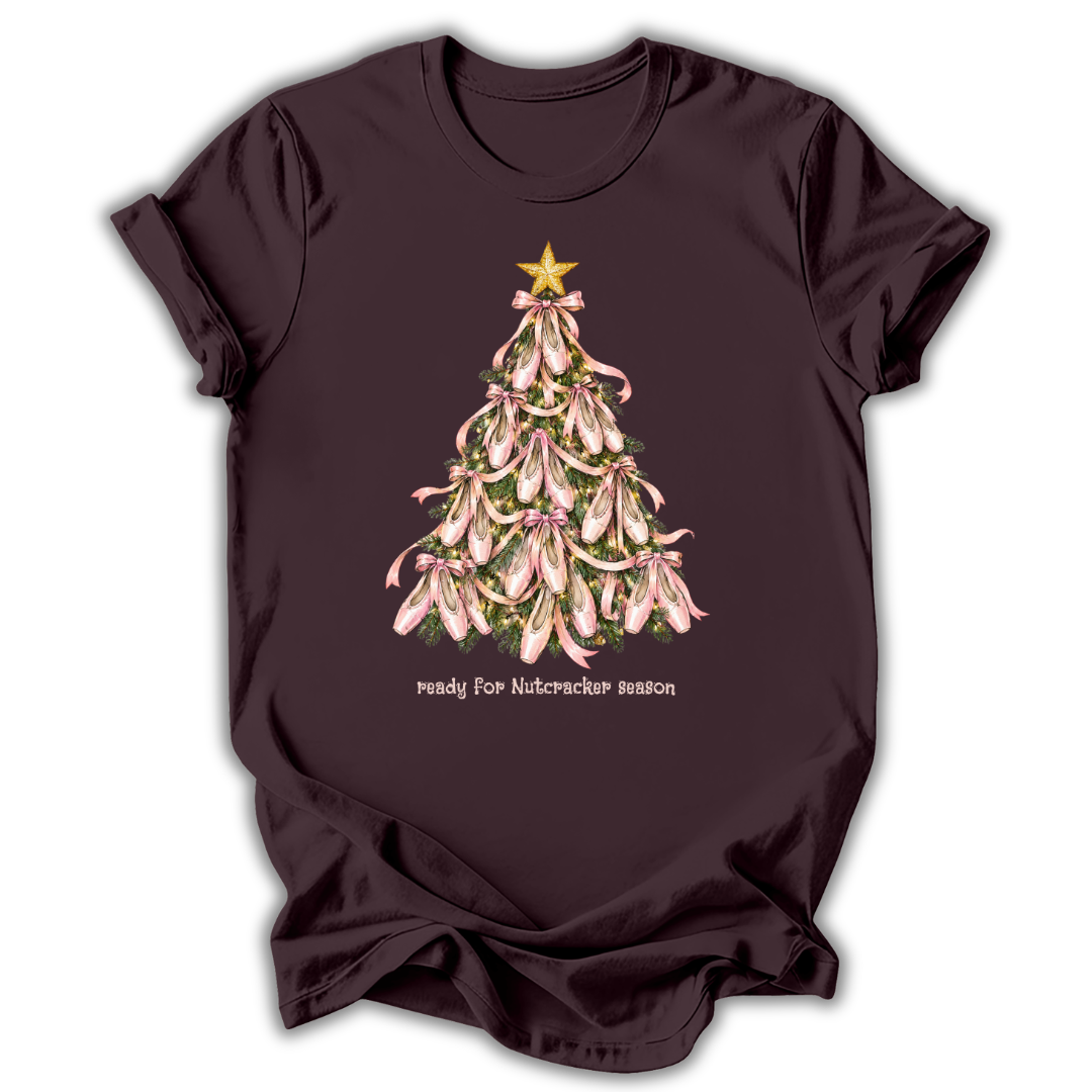 A dark plum-colored short-sleeve t-shirt with a graphic of a Christmas tree made of pink lilies with a gold star on top, and the text 'ready for Nutcracker season' printed below.