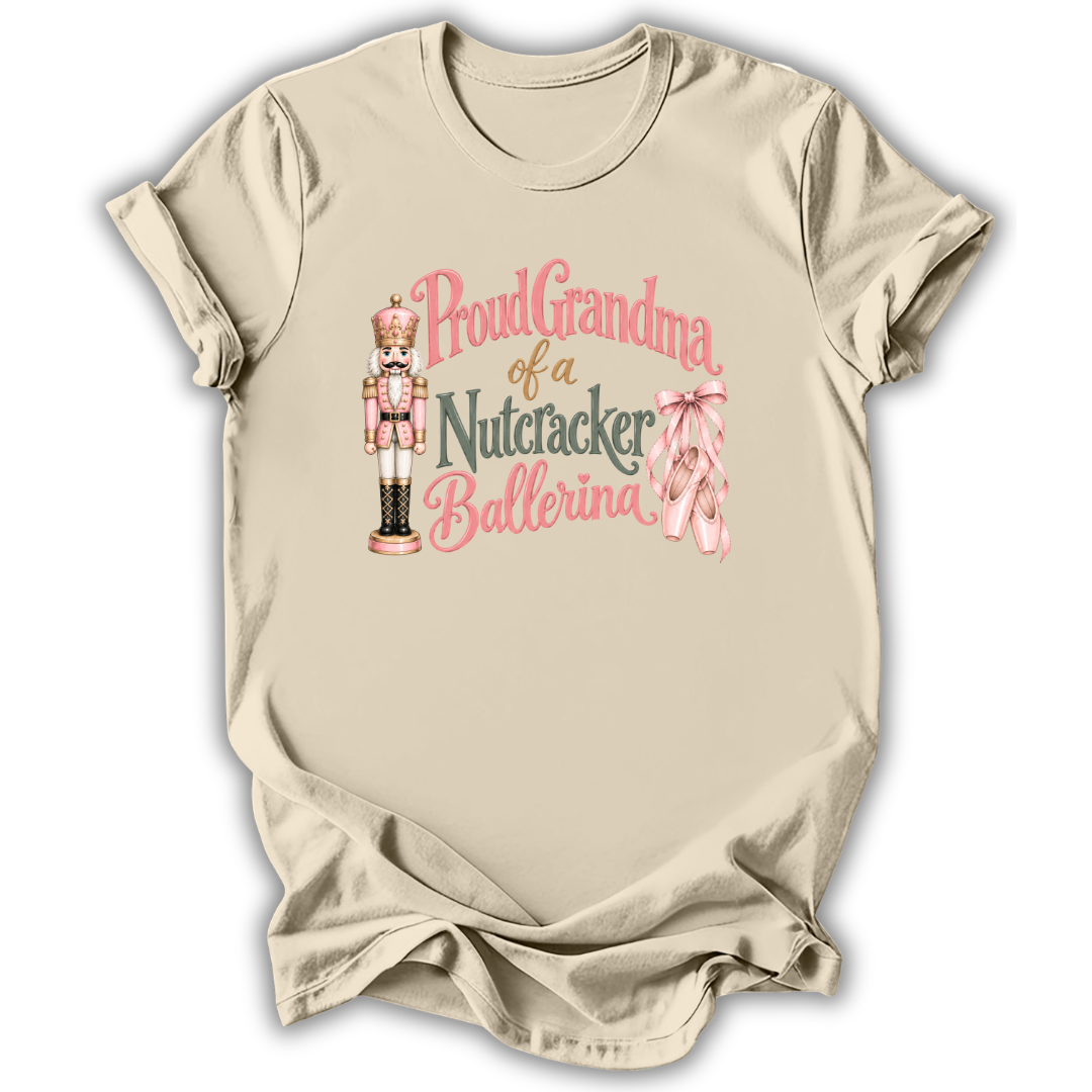 A beige short-sleeve crewneck t-shirt with a graphic print that reads 'Proud Grandma of a Nutcracker Ballerina' in pink and dark gray lettering, accompanied by an illustration of a nutcracker and pink ballet slippers.