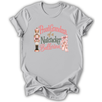 A light gray short-sleeve t-shirt with a graphic on the front that reads 'Proud Grandma of a Nutcracker Ballerina' in pink and dark green lettering, accompanied by an illustration of a nutcracker and pink ballet slippers.