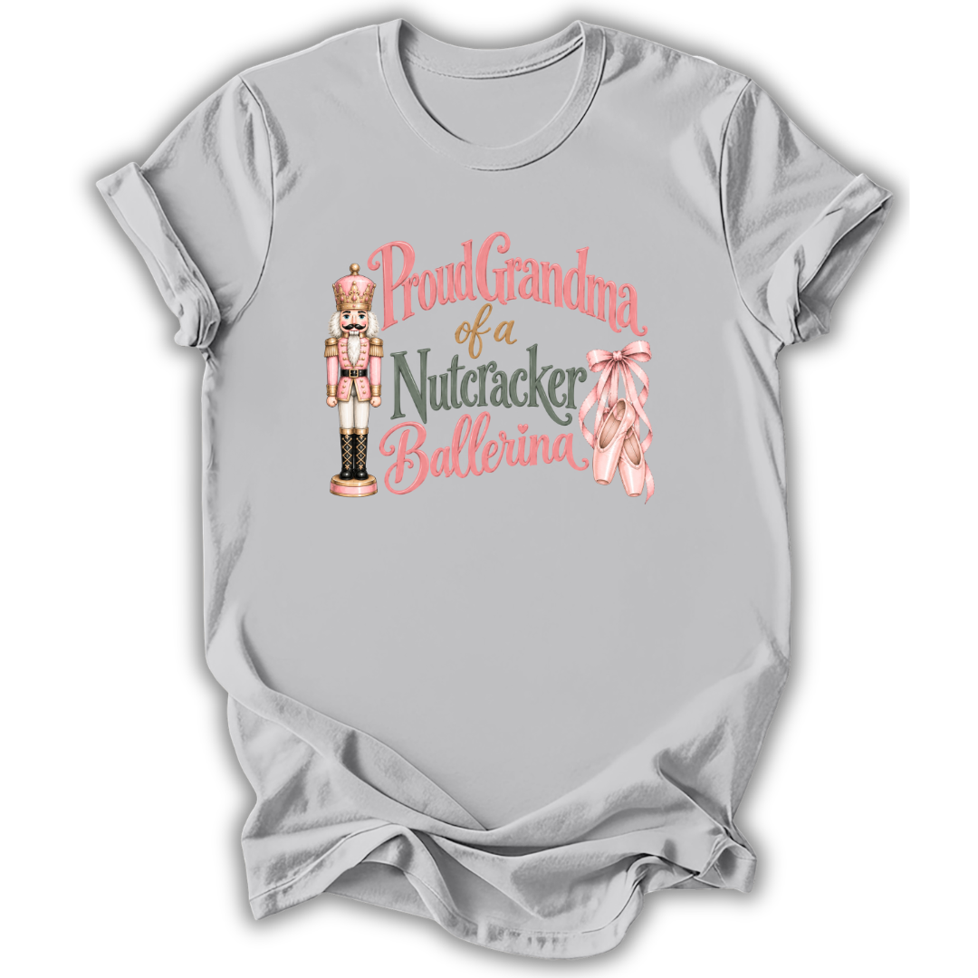 A light gray short-sleeve t-shirt with a graphic on the front that reads 'Proud Grandma of a Nutcracker Ballerina' in pink and dark green lettering, accompanied by an illustration of a nutcracker and pink ballet slippers.