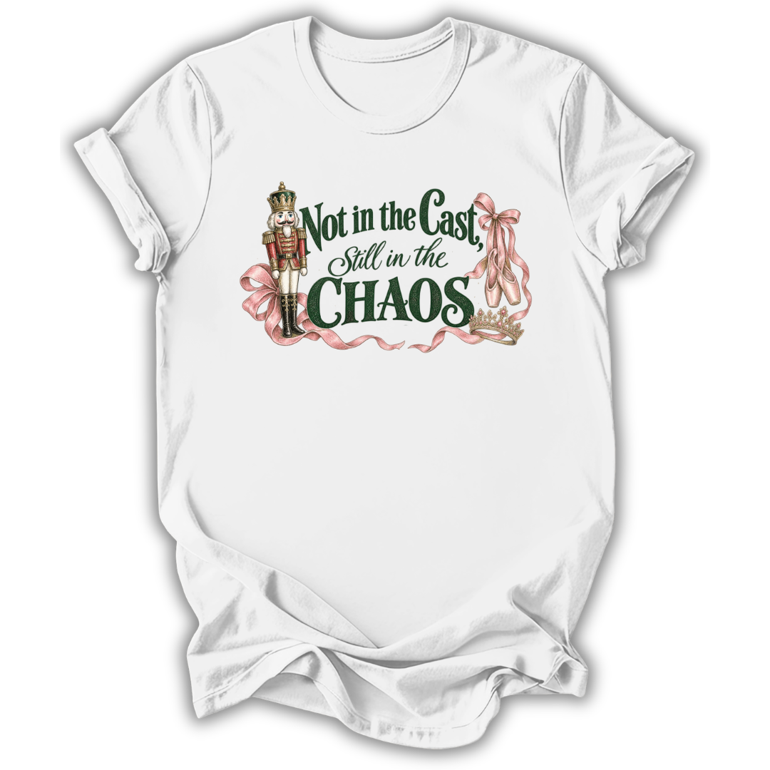 A white short-sleeve t-shirt with a graphic print on the front. The graphic features a nutcracker soldier, pink ribbons, and the text 'Not in the Cast, Still in the CHAOS' in dark green and pink lettering.