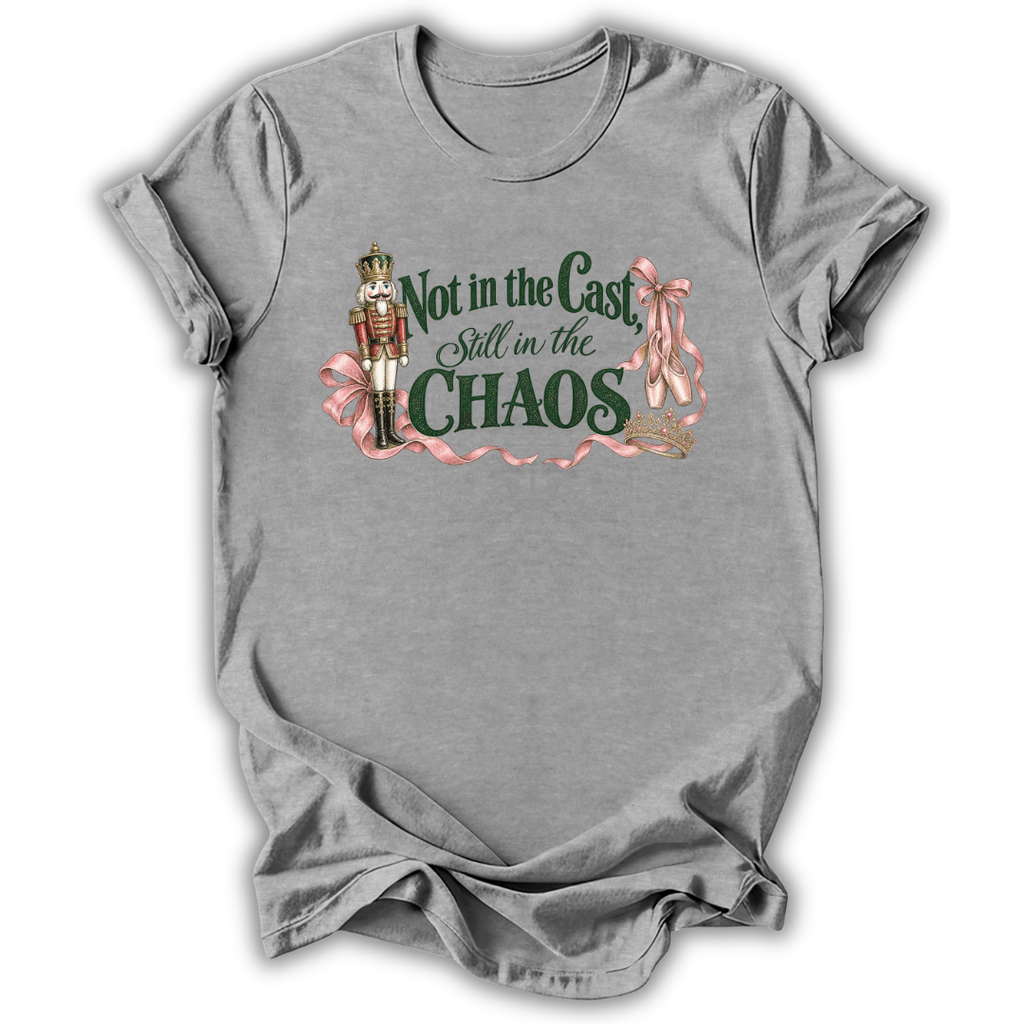 A heather gray short-sleeve t-shirt with a graphic print on the front. The graphic features a nutcracker soldier, pink ribbons, and the text 'Not in the Cast Still in the CHAOS' in dark green and pink lettering.