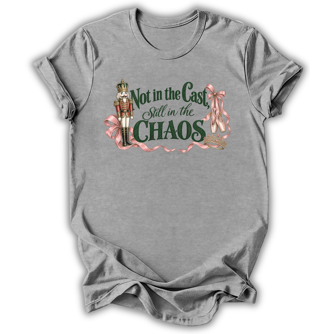 A heather gray short-sleeve t-shirt with a graphic print on the front. The graphic features a nutcracker soldier, pink ribbons, and the text 'Not in the Cast Still in the CHAOS' in dark green and pink lettering.