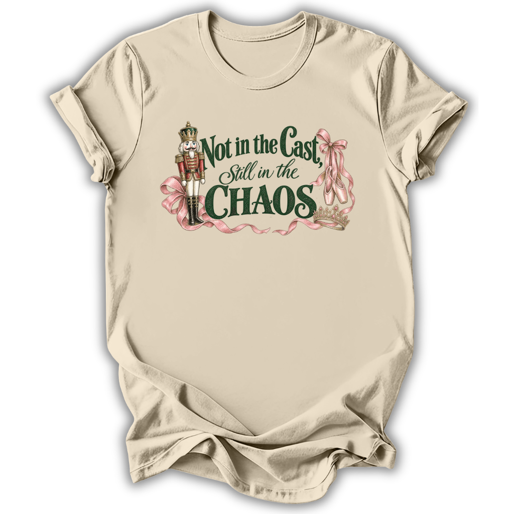 A beige short-sleeve crewneck t-shirt with a graphic print on the front. The graphic features a nutcracker soldier, pink ribbons, and the text 'Not in the Cast, Still in the CHAOS' in dark green and pink lettering.