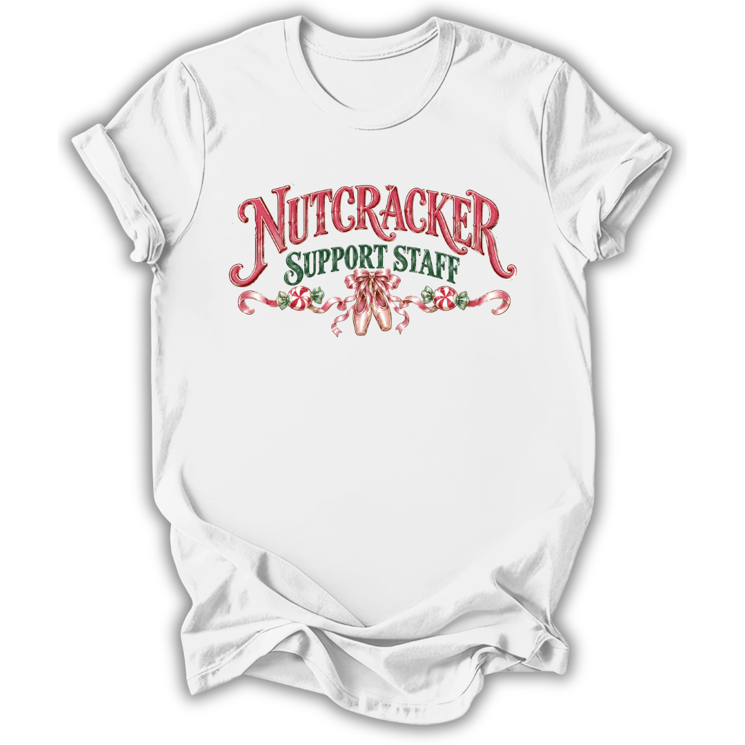 A white short-sleeve crewneck t-shirt with a festive graphic on the chest reading 'NUTCRACKER SUPPORT STAFF' in red and green lettering, decorated with candy canes and a nutcracker silhouette.