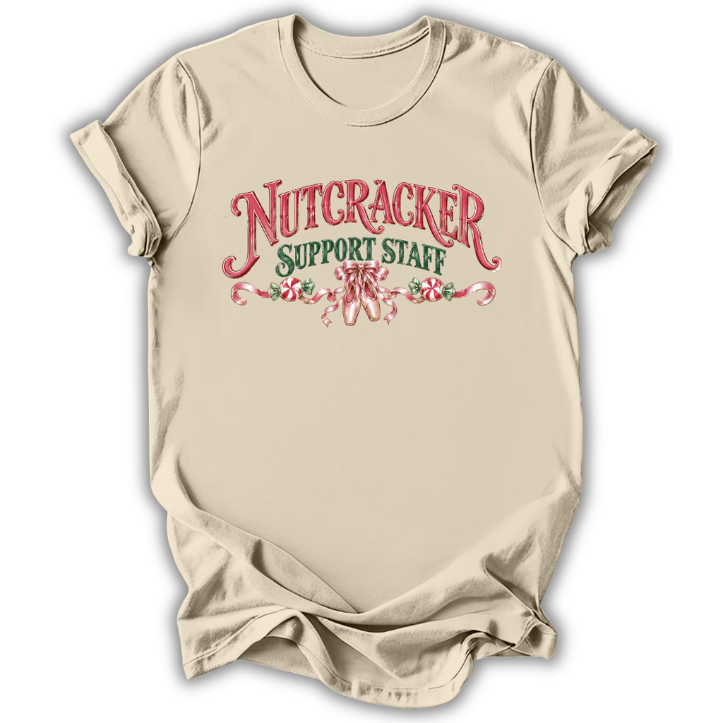 A beige short-sleeve crewneck t-shirt with a graphic print on the front reading 'NUTCRACKER SUPPORT STAFF' in red and green lettering with candy cane illustrations, displayed on a white background.