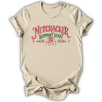 A beige short-sleeve crewneck t-shirt with a graphic print on the front reading 'NUTCRACKER SUPPORT STAFF' in red and green lettering with candy cane illustrations, displayed on a white background.