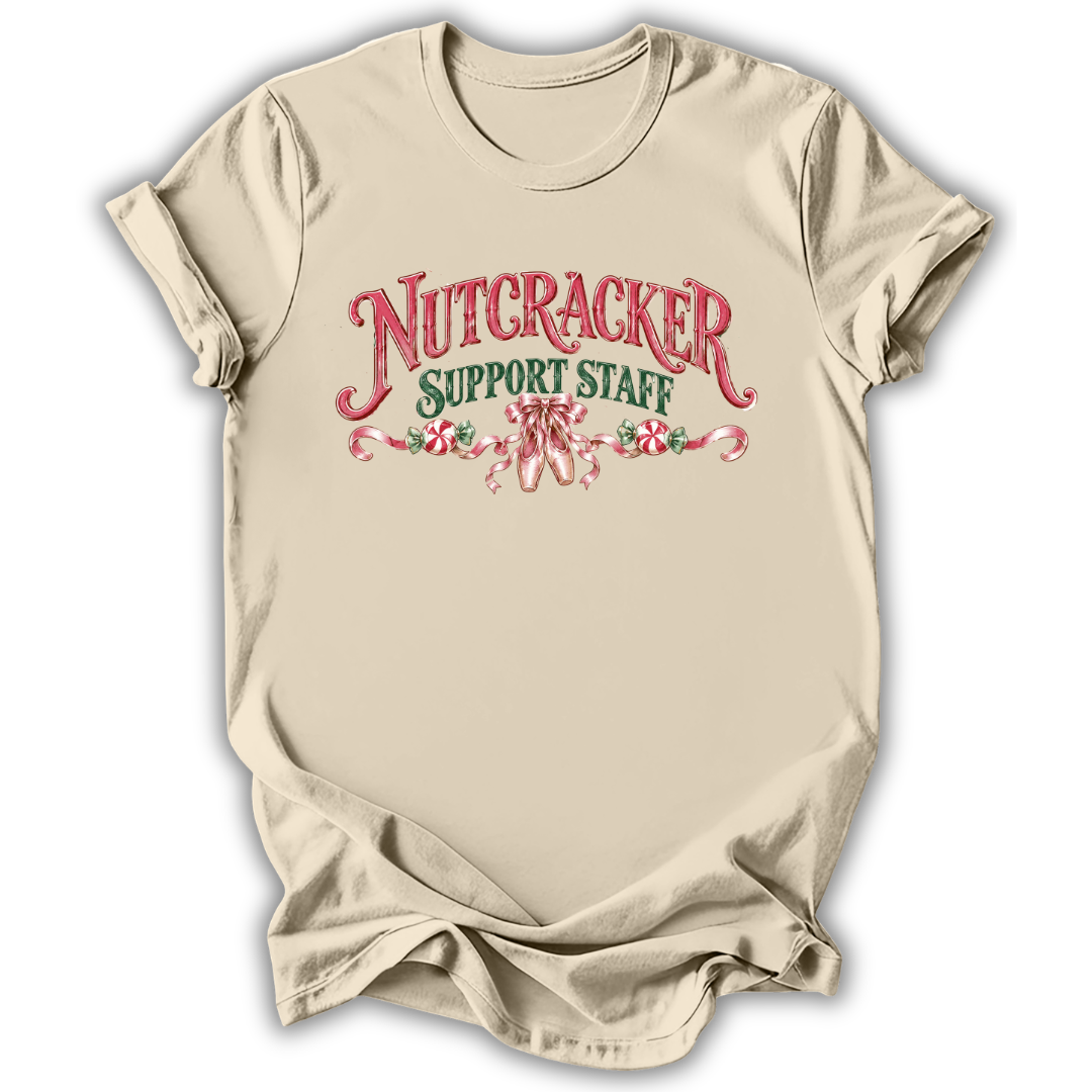 A beige short-sleeve crewneck t-shirt with a graphic print on the front reading 'NUTCRACKER SUPPORT STAFF' in red and green lettering with candy cane illustrations, displayed on a white background.