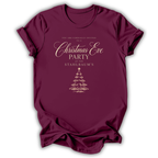 A maroon short-sleeve t-shirt with a gold printed Christmas Eve party invitation design on the front, displayed against a clean white background.