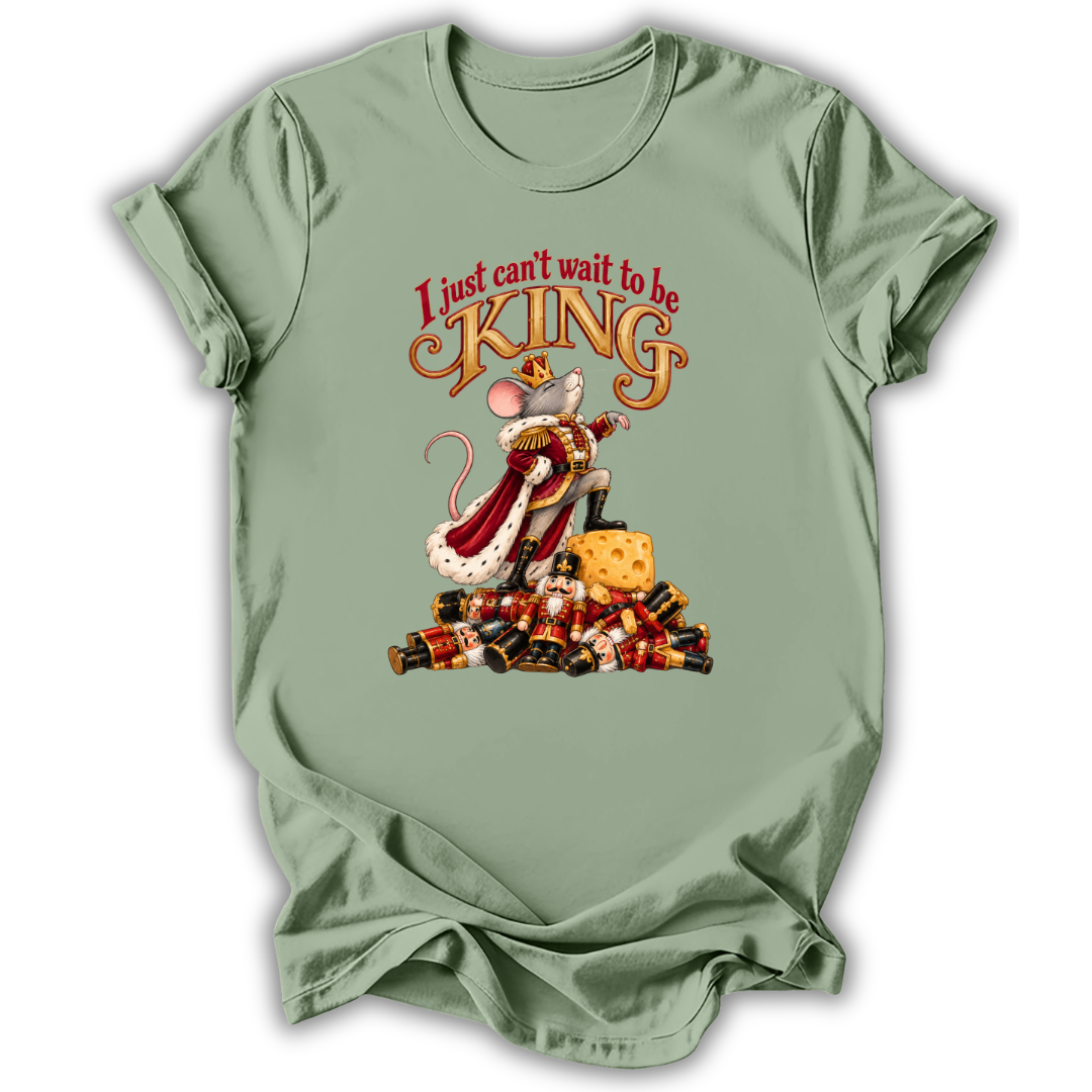 A sage green short-sleeve t-shirt with a graphic of a mouse dressed as a king sitting on a pile of gold coins, with the text 'I just can't wait to be KING' above it.