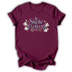 A maroon short-sleeve t-shirt with a graphic on the chest that reads 'SNOW FLOWERS REPEAT' in blue and gold lettering, decorated with white snowflakes and flowers, shown on a white background.