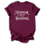 A maroon short-sleeve crewneck t-shirt with the phrase 'Nutcracker Magic Starts Backstage' printed in a cream-colored decorative font on the front, displayed against a clean white background.
