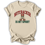 A beige short-sleeve t-shirt with a graphic print that reads 'NUTCRACKER IS MY SPORT' in red and dark green lettering, featuring a nutcracker illustration in the center, shown on a white background with a soft drop shadow.
