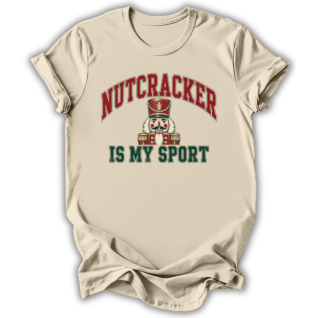 A beige short-sleeve t-shirt with a graphic print that reads 'NUTCRACKER IS MY SPORT' in red and dark green lettering, featuring a nutcracker illustration in the center, shown on a white background with a soft drop shadow.