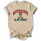 A beige short-sleeve t-shirt with a graphic print on the front. The graphic features the word 'NUTCRACKER' in red arched letters above a nutcracker illustration, and the phrase 'IS MY SPORT' in dark green block letters below.
