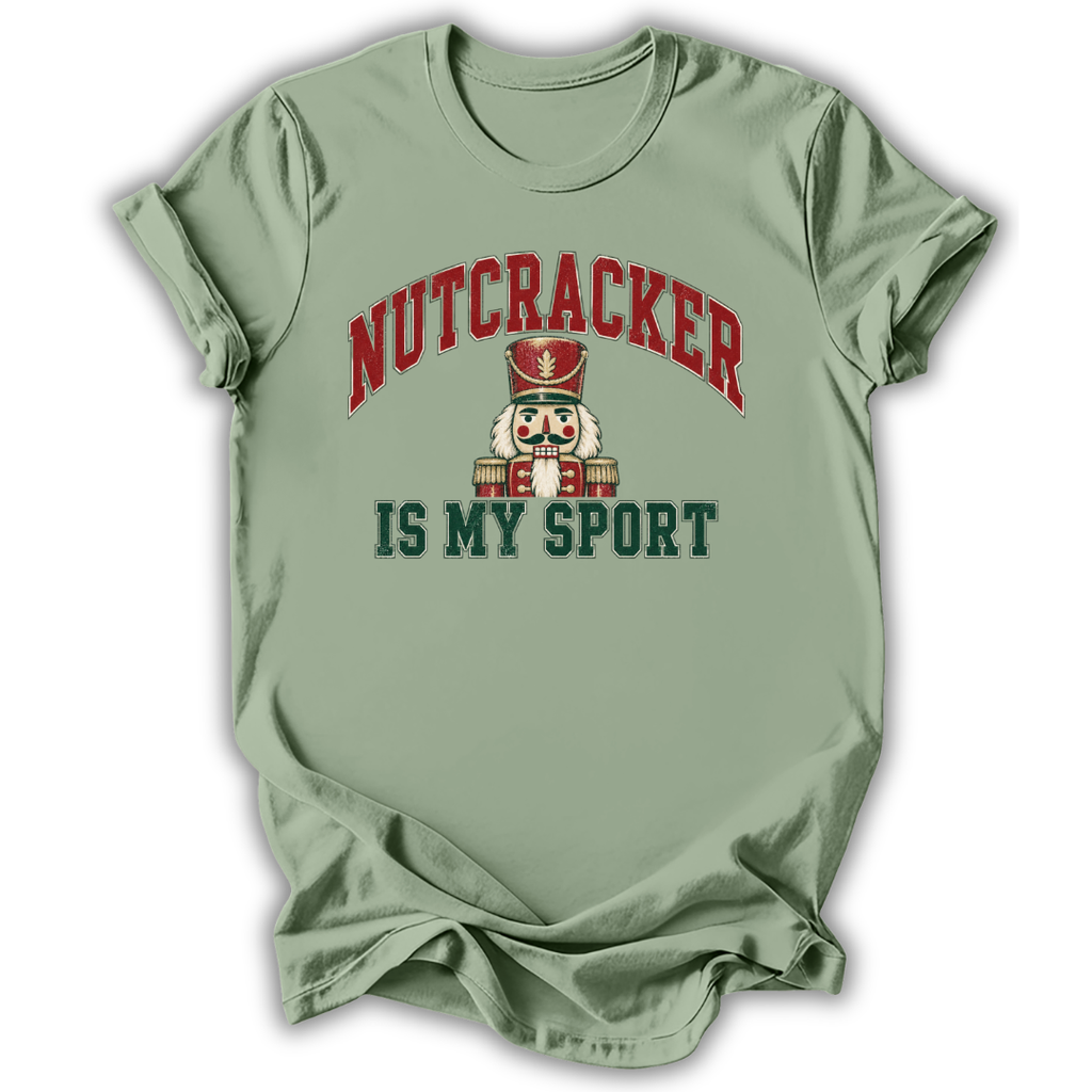 A sage green short-sleeve t-shirt with a graphic print on the front. The print features the word 'NUTCRACKER' in red arched letters above a nutcracker illustration, and the phrase 'IS MY SPORT' in dark green block letters below.