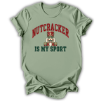 A sage green short-sleeve t-shirt with a graphic print on the front. The print features the word 'NUTCRACKER' in red arched letters above a nutcracker illustration, and the phrase 'IS MY SPORT' in dark green block letters below.