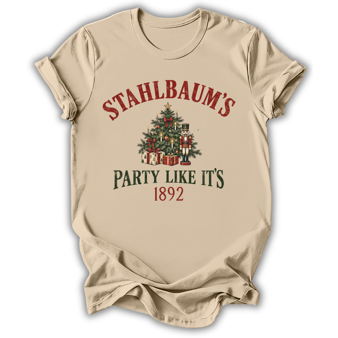 A beige short-sleeve t-shirt with a vintage-style Christmas graphic on the front. The graphic features the text 'STAHLBAUM'S PARTY LIKE IT'S 1892' in red and dark green lettering, surrounding an illustration of a decorated Christmas tree, gift boxes, and nutcrackers.