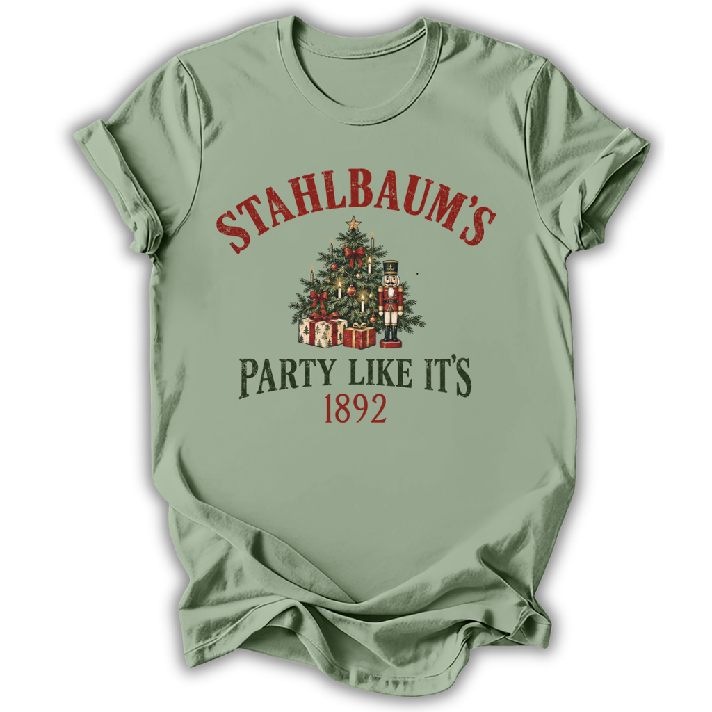 A sage green short-sleeve t-shirt with a vintage Christmas graphic on the front. The graphic features the text 'STAHLBAUM'S PARTY LIKE IT'S 1892' in red and dark green lettering, surrounding an illustration of a decorated Christmas tree, gift boxes, and nutcrackers.