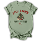 A sage green short-sleeve t-shirt with a vintage Christmas graphic on the front. The graphic features the text 'STAHLBAUM'S PARTY LIKE IT'S 1892' in red and dark green lettering, surrounding an illustration of a decorated Christmas tree, gift boxes, and nutcrackers.