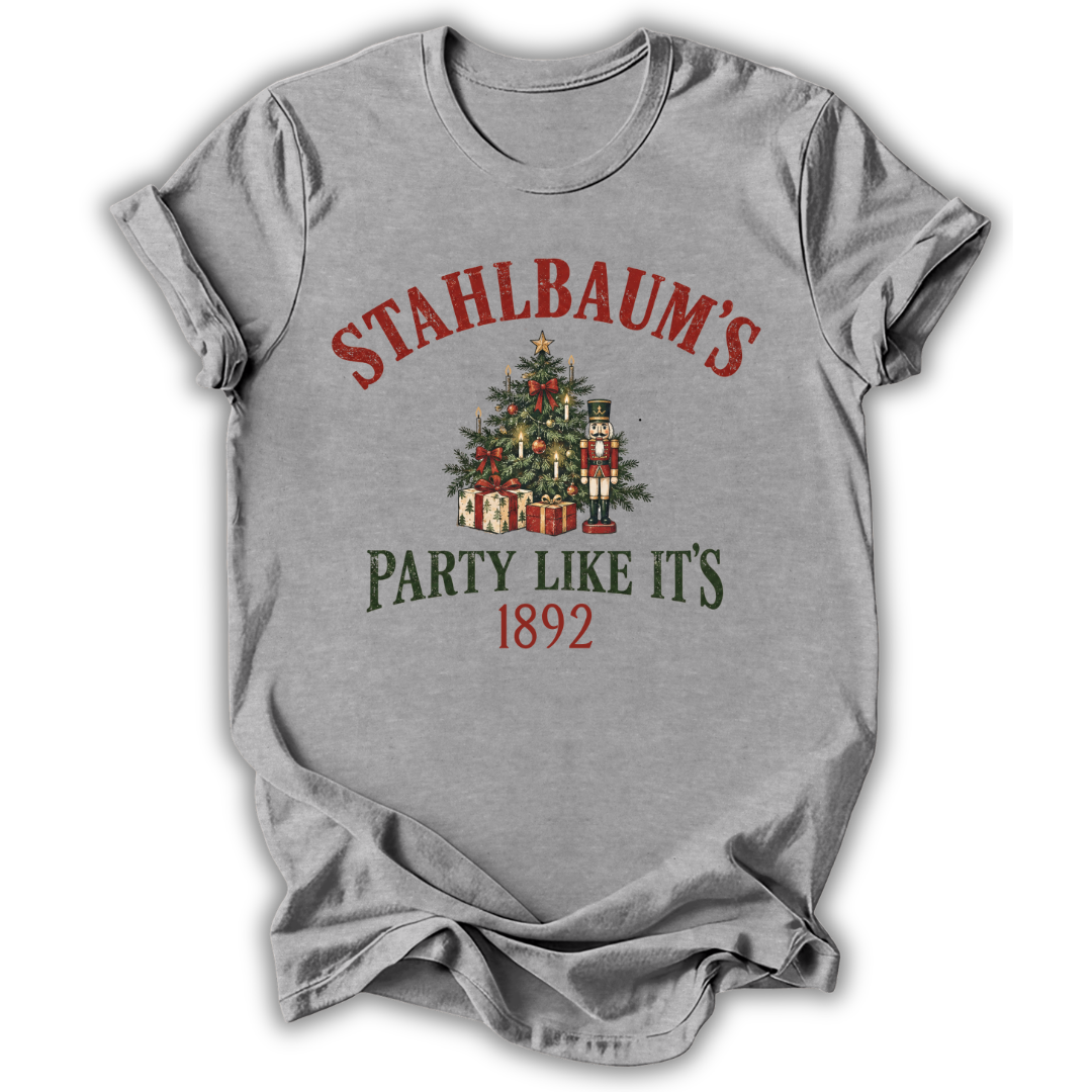 A heather gray short-sleeve t-shirt with a vintage-style Christmas graphic on the front. The graphic features the text 'STAHLBAUM'S PARTY LIKE IT'S 1892' in red and dark green lettering, surrounding an illustration of a decorated Christmas tree, gifts, and nutcrackers.