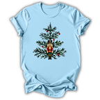 A light blue short-sleeve crewneck t-shirt with a graphic of a nutcracker standing in front of a decorated Christmas tree, shown on a plain white background.