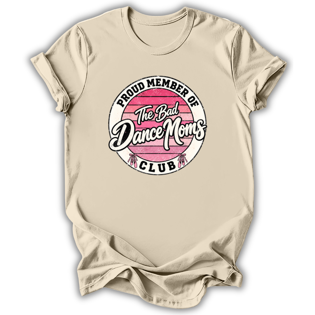 A beige short-sleeve t-shirt with a circular graphic on the chest that reads 'PROUD MEMBER OF The Bad Dance Moms CLUB'. The graphic features a pink striped circle with white text and small pink and white dance figures at the bottom.