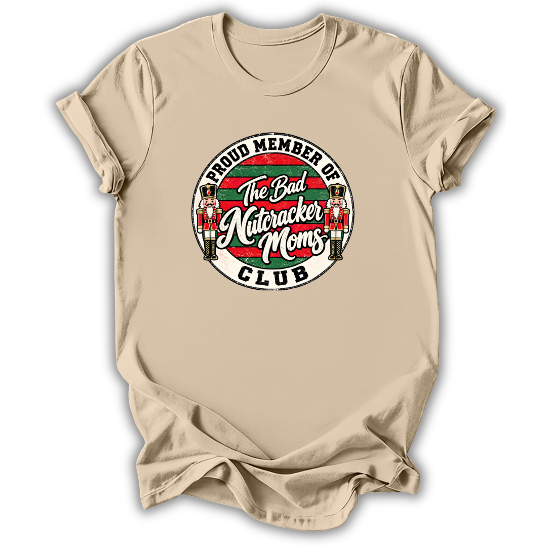 A beige short-sleeve t-shirt with a circular graphic on the chest that reads 'PROUD MEMBER OF The Bad Nutcracker Moms CLUB' with nutcracker figures and red, green, and white stripes.