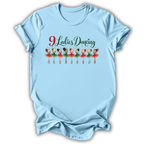 A light blue short-sleeve t-shirt with a graphic on the chest that reads '9 Ladies Dancing' above a row of illustrated ballerinas in red and green tutus, shown on a plain white background.