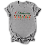 A heather gray short-sleeve t-shirt with a graphic print on the front that reads '9 Ladies Dancing' in green and red script above a row of nine cartoon ballerinas in red and green outfits, shown on a plain white background.