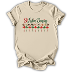 A beige short-sleeve t-shirt with a graphic print on the front that reads '9 Ladies Dancing' in red and green script above a row of nine cartoon ballerinas in red and green tutus, shown on a white background.