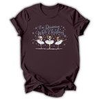 Brown short-sleeve t-shirt with a graphic print of three ballerinas and the text 'I'm Dreaming While Christmas' in blue and white, displayed on a clean white background.