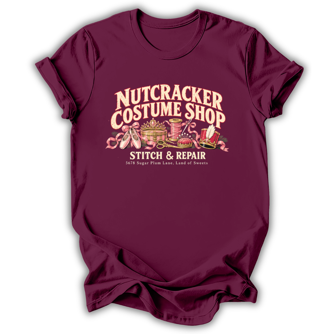 A maroon short-sleeve t-shirt printed with the “Nutcracker Costume Shop” graphic design.