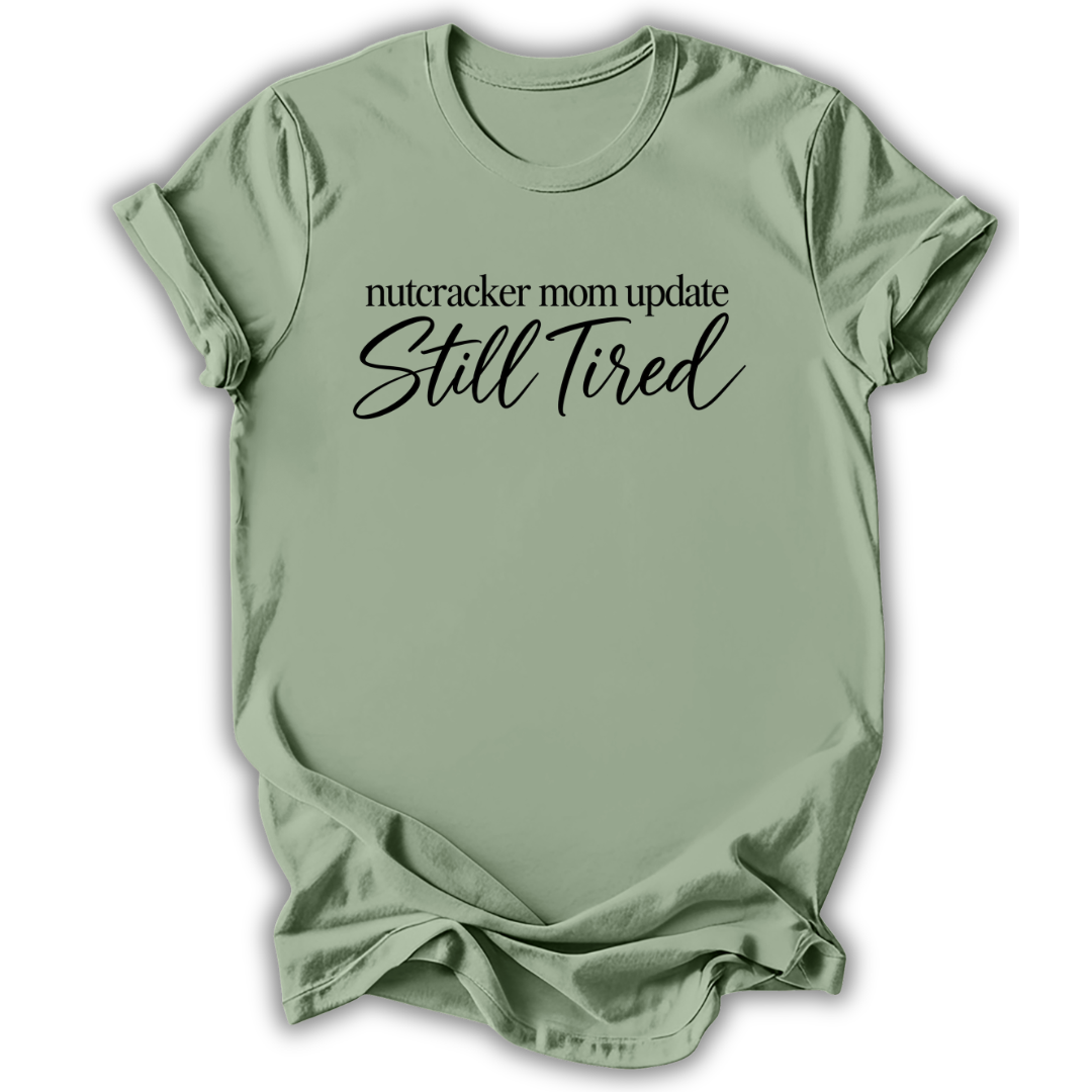 A sage green short-sleeve t-shirt printed with the “Still Tired Nutcracker Mom” graphic design.