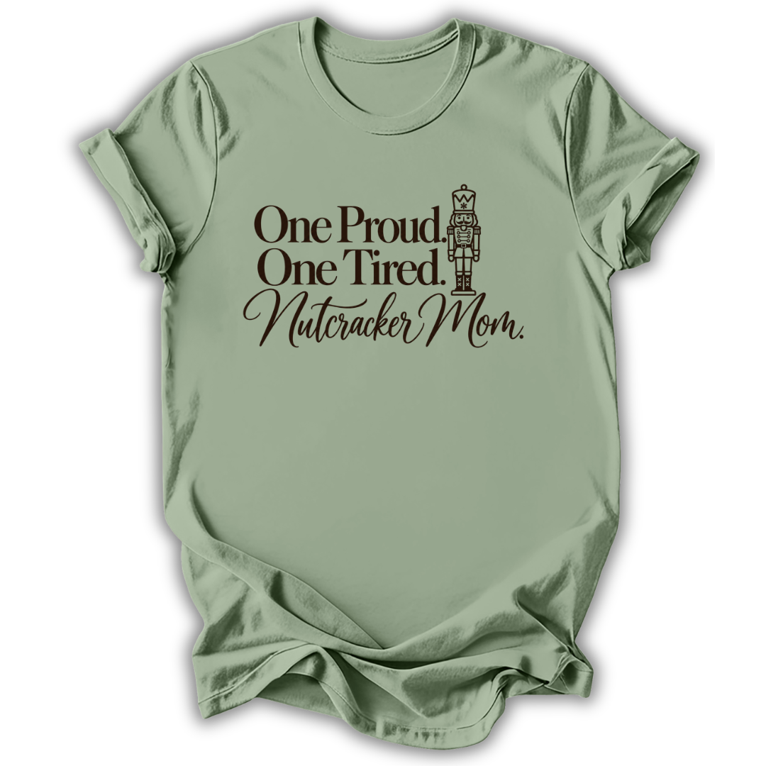 A sage green short-sleeve t-shirt printed with the “Proud & Tired Nutcracker Mom” graphic design.