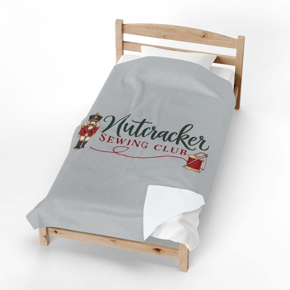 A light gray bedding set, including a comforter and a matching pillowcase, displayed on a wooden toddler bed. The comforter features a printed graphic of a nutcracker soldier and the text 'Nutcracker Sewing Club' in dark green and red script, with a small red spool of thread illustration.