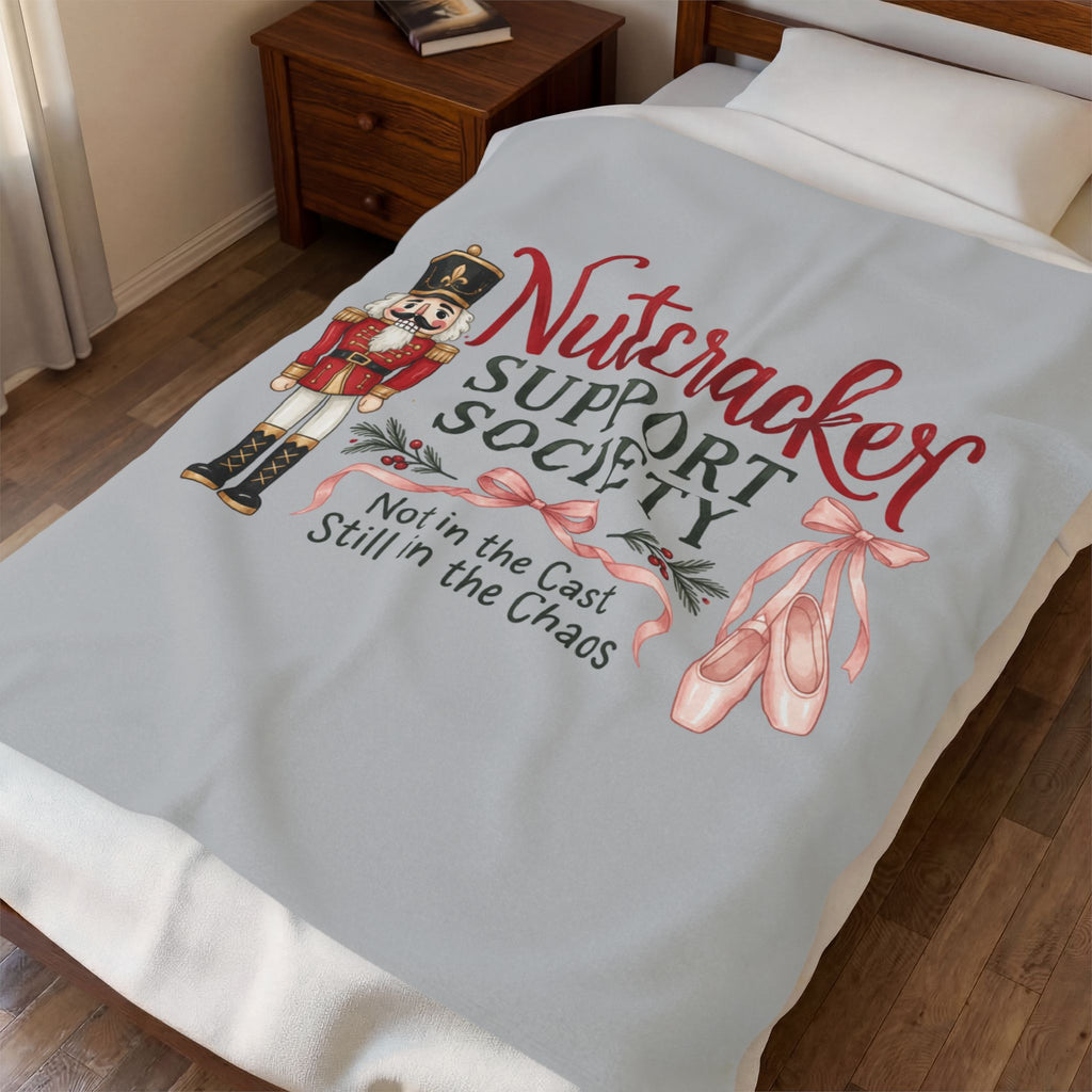 A light gray fleece blanket with a colorful 'Nutcracker Support Society' graphic is draped over a bed in a bedroom setting.