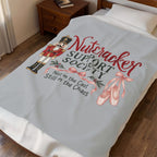 A light gray fleece blanket with a colorful 'Nutcracker Support Society' graphic is draped over a bed in a bedroom setting.