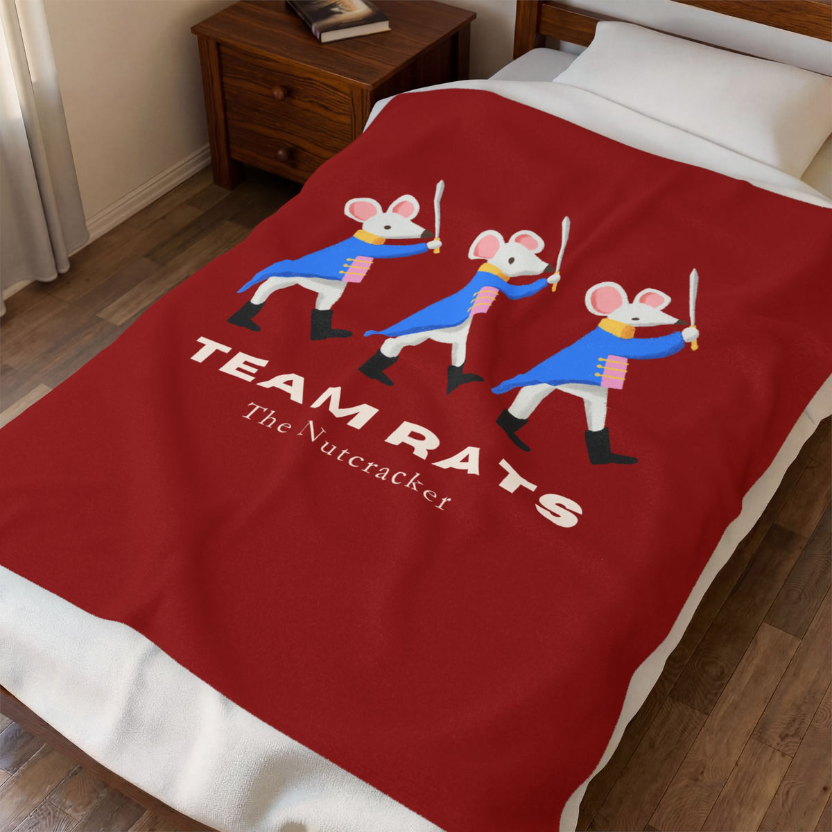 A red blanket with a cartoon graphic of three rats in blue and white uniforms holding swords, with the text 'TEAM RATS' and 'The Nutcracker', shown on a bed in a bedroom setting.
