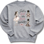 A heather gray crewneck sweatshirt featuring a graphic print of a ballerina in a pink dress, a nutcracker, a cat, and the text 'Just a Girl Who Loves Nutcracker' in pink and black script.